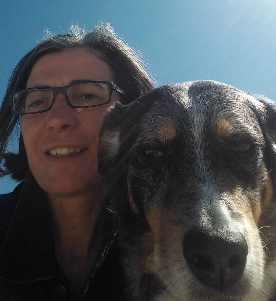 frau mit brille vor blauem Sonnenhimmel mit weiss, schwarz, braunem Hund im Vordergrund